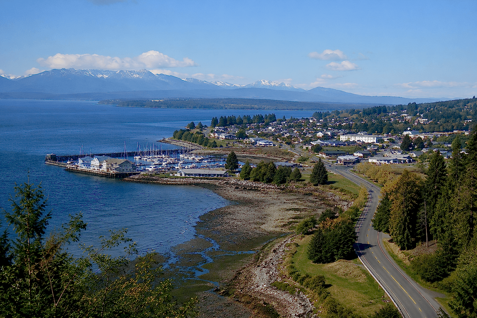 oak-harbor-wa-waterfront-marina-aerial-view Aerial view of Oak Harbor waterfront marina with coastal shoreline, scenic highway, evergreen trees, and mountain views on Whidbey Island